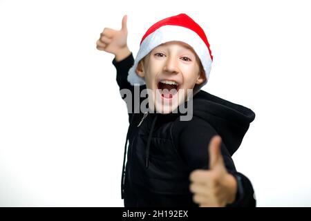 Ein Junge in einem roten Weihnachtsmann-Hut, der auf weißem Hintergrund steht, gibt den Daumen nach oben und lacht laut Stockfoto