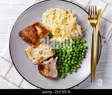 Gebratenes Schweinelende und Sauerkraut auf einem Teller mit grünen Erbsen auf einem weißen Holztisch mit Besteck, Draufsicht, Nahaufnahme Stockfoto