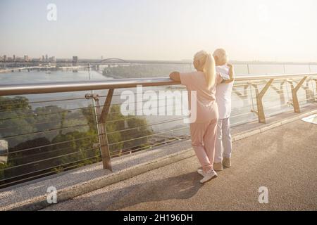 Die ältere Dame umarmt ihren Mann in Sportkleidung und genießt den Blick auf die Stadt von der Fußgängerbrücke Stockfoto