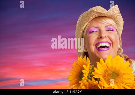 Schönes lachendes Mädchen, das Cowboy-Hut mit gelben Sonnenblumen gegen lila und rosa Sonnenuntergang trägt Stockfoto