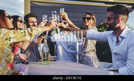 Lachende Freunde toasten Weingläser, während sie zusammen sitzen. Fröhliche Gruppe von Freunden, die vor einem Luxushotel feiern. Zwei glückliche Paare genießen Stockfoto