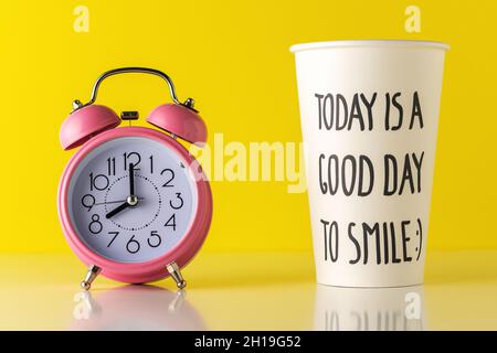 Rosafarbener Wecker mit einer Papiertasse Kaffee auf einem Holzschreibtisch. Minimaler Zeitbegriff. Stockfoto