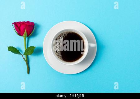 Kreative Komposition aus roter Rosenblüte und Tasse Kaffee auf blauem Hintergrund. Minimales Federkonzept. Stockfoto