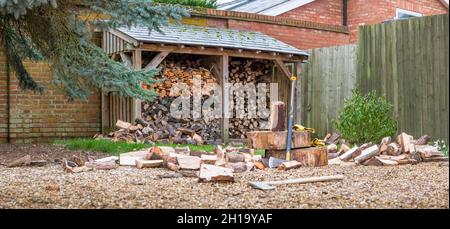 Holzstapel mit Axt und spaltenden Maul. Britische Gartenpanoramaszene, die das Konzept erneuerbarer Energien und Biokraftstoffe darstellt Stockfoto