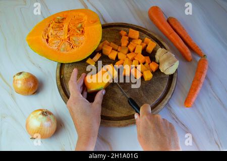 Herbstliche Gerichte, Kürbissuppenkochen.Weibliche Hände schneiden einen Kürbis auf einem Holzbrett, Zwiebeln, Ingwer und Karotten liegen in der Nähe Stockfoto