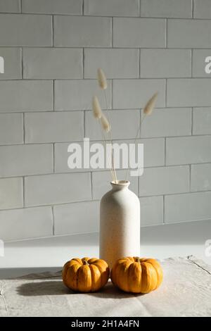 Stillleben im Herbst. Orangefarbene Kürbisse und Vase mit trockenen Blumen auf textiler Serviette. Weiße Backsteinmauer auf dem Hintergrund. Thanksgiving-Tagesdekor, Skandinavien Stockfoto