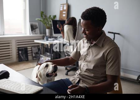 Porträt eines lächelnden afroamerikanischen Mannes, der während der Arbeit im Büro einen Hund streichelt, haustierfreundlicher Arbeitsplatz Stockfoto