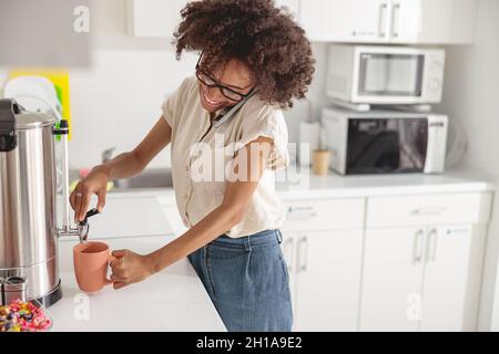 Glückliche Dame, die Tee in eine Tasse gießt und in der Küche telefoniert Stockfoto