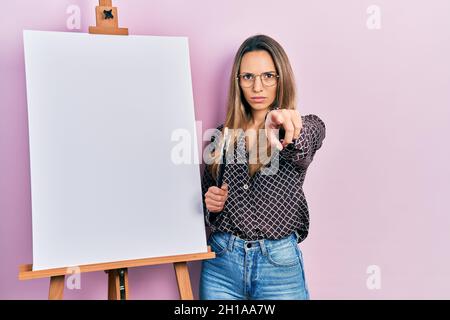 Schöne hispanische Frau, die beim Maler Staffelstand steht und Bürsten hält, die mit dem Finger auf die Kamera und auf Sie zeigen, selbstbewusste Geste, die seri zeigt Stockfoto