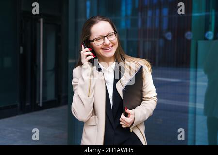 Geschäftsfrau aus dem Kaukasus in Anzug und Brille mit Laptop in der Hand, die in der Nähe des Business Centers telefoniert. Konzept einer stilvollen und unabhängigen Frau an Wochentagen vor dem Bürogebäude Stockfoto