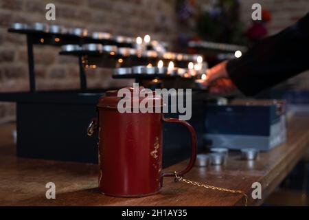 Hand zündet Kerze in der Kirche an, nachdem sie Geld für Spenden in die rote Metallkassebox geworfen hat. Konzentrieren Sie sich auf den Rand des Sammelkastens auf dem Tisch Stockfoto