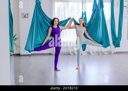 Konzentrierte junge Frauen, die Antigravity Yoga mit Hängematte mit Hilfe des persönlichen Trainers tun Stockfoto