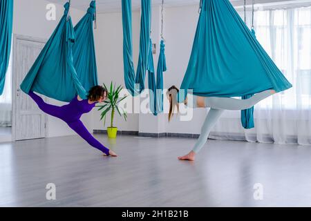 Konzentrierte junge Frauen, die Antigravity Yoga mit Hängematte mit Hilfe des persönlichen Trainers tun Stockfoto