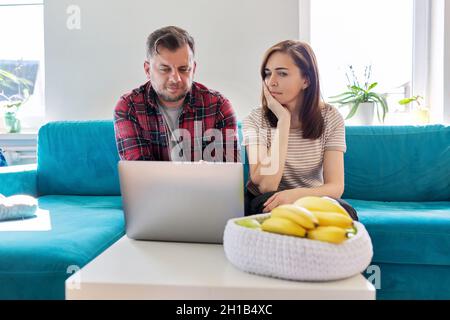 Ernsthaftes Paar mittleren Alters, das zu Hause im Wohnzimmer einen Laptop-Bildschirm ansieht Stockfoto