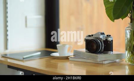 Nahaufnahme tragbarer Arbeitsplatz im Café mit Vintage-Kamera auf Büchern, Kaffeetasse und digitalem Tablet auf Holztisch. Stockfoto