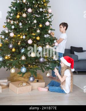 Kinder - ein Junge und ein Mädchen spielen in der Nähe des Weihnachtsbaums. Wohnzimmer mit Weihnachtsbaum und Dekorationen. Neujahr. Geschenk geben. Stockfoto