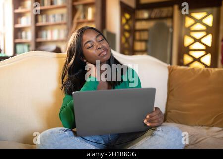 Müde junge schwarze Frau, die zu Hause auf dem Laptop auf der Couch arbeitet, den Hals massiert, Kopfschmerzen leidet und sich erschöpft fühlt Stockfoto