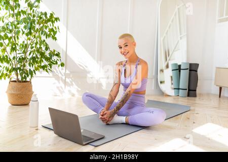 Inländisches Yoga-Konzept. Junge, fittierte Dame macht eine Schmetterlingsstraffungsübung und meditiert zu Hause vor dem Laptop Stockfoto