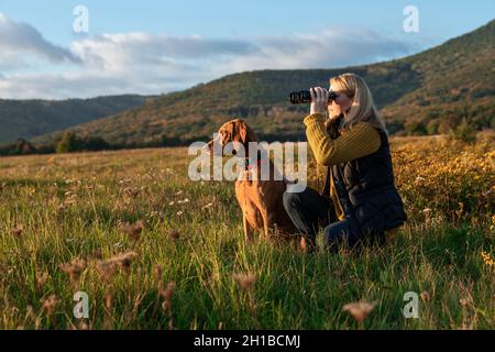 Die junge Jägerin, die ein Fernglas verwendet, um Vögel mit dem ungarischen Vizsla-Hund an ihrer Seite zu beobachten, an einem schönen sonnigen Herbstabend auf einer Wiese. Hun Stockfoto