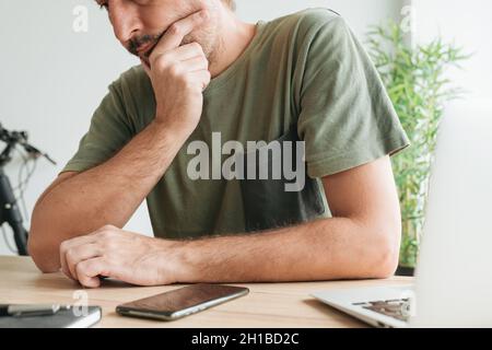 Telearbeiter, der am Schreibtisch des Heimbüros sitzt und mit der Hand am Kinn über Remote-Arbeit denkt, selektiver Fokus Stockfoto