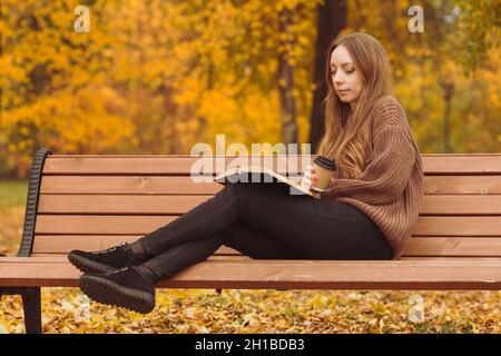Die junge Frau in einem Herbstpark sitzt auf einer Bank, trinkt Kaffee und liest ein Buch. Konzept - Menschen im Herbst. Stockfoto
