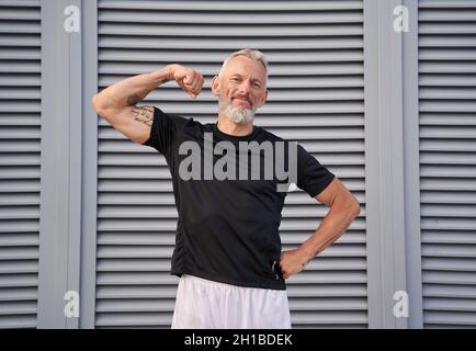 Sportlich reifer Mann, der die Kamera anschaut, seinen muskulösen Arm und seinen Bizeps zeigt, während er draußen auf grauem Hintergrund steht Stockfoto