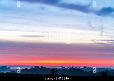 Weitwinkelansicht eines fernen dramatischen rot-gelben Himmels über der nebelbedeckten Landschaft in Kent, England. Niedriger Horizont mit einem Band aus rotem und gelbem Himmel und über diesem blauen Himmel in der frühen Morgendämmerung Stockfoto