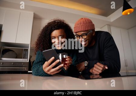 Multiethnisches Paar, das in einer modernen Wohnung in der Küche am Telefon lächelt Stockfoto