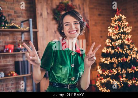 Foto von entzückenden glänzenden jungen Frau gekleidet grünen Kostüm lächelnd feiert Neujahr zeigt V-Zeichen drinnen Zimmer Haus Stockfoto