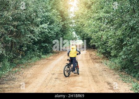 Kleiner Junge in gelber Weste fährt im Sommer auf einem modernen Fahrrad auf einer ländlichen Landstraße mit grünen Bäumen an den Seiten Stockfoto