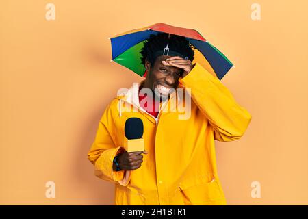 Junger afroamerikanischer Journalist mit gelbem Regenmantel und Regenschirmkappe gestresst und frustriert mit der Hand am Kopf, überrascht und wütend im Gesicht Stockfoto