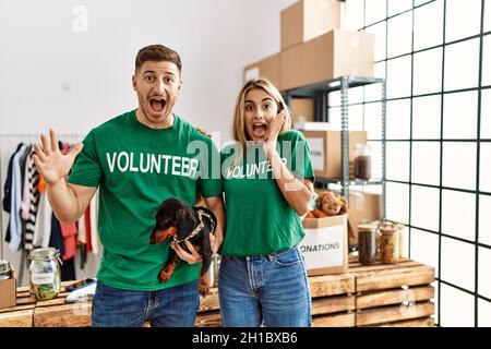 Junges Paar mit niedlichem Hund, das ein T-Shirt mit Freiwilligen trägt, am Spendenstand, um den Sieg mit einem glücklichen Lächeln und einem Siegerausdruck mit erhobenen Händen zu feiern Stockfoto