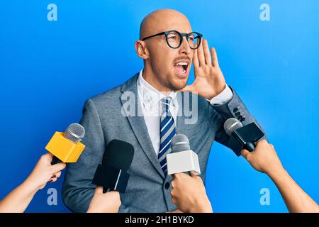 Kahlköpfiger Mann mit Bart, der von Reportern interviewt wird, die Mikrofone mit Schreien und Schreien laut an der Seite halten, mit der Hand auf dem Mund. Kommunikationskonzept. Stockfoto