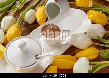 Von Hand Milch in den Kaffee in der Nähe von weißen und gelben Tulpen auf Holztisch Draufsicht gießen. Romantisches Frühstückskonzept Stockfoto