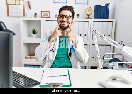 Junger Mann mit Bart, der eine Arztuniform und ein Stethoskop in der Klinik trug, lächelte mit offenem Mund, die Finger zeigten und ein fröhliches Lächeln erzwangen Stockfoto