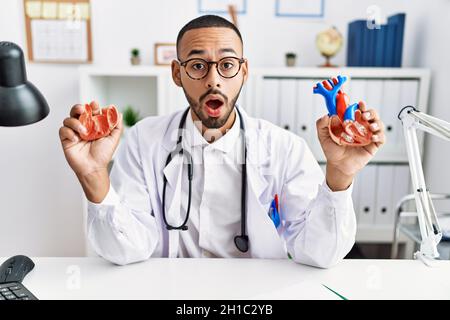 afroamerikanischer Arzt, der anatomisches Herz in der Klinik im Schockgesicht hielt, skeptisch und sarkastisch aussachte, überrascht mit offenem Mund Stockfoto