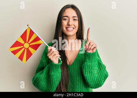 Junge hispanische Mädchen hält mazedonische Flagge lächelnd mit einer Idee oder Frage Zeigefinger mit glücklichen Gesicht, Nummer eins Stockfoto