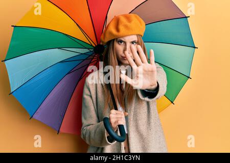 Junge irische Frau im französischen Stil hält bunten Regenschirm mit offener Hand tun Stop-Zeichen mit ernst und selbstbewusst Ausdruck, Verteidigung Geste Stockfoto