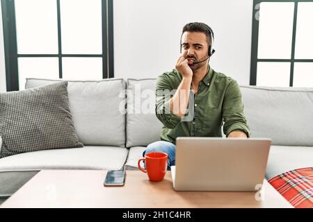 Junger hispanischer Mann mit Bart, der ein Callcenter-Headset trägt, der von zu Hause aus gestresst und nervös aussieht, mit Händen auf dem Mund, beißende Nägel. Angst Stockfoto