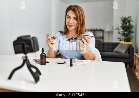 Kaukasische Frau mittleren Alters macht Make-up-Tutorial mit Kamera sitzen auf dem Tisch zu Hause. Stockfoto