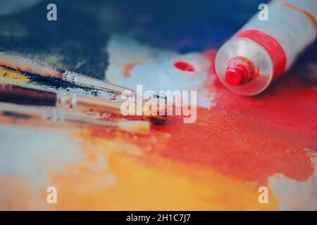 Auf der Palette, die mit verschiedenen leuchtenden Farben bemalt ist, befindet sich ein Rohr aus rosa künstlerischem Öl und drei Pinsel. Der Prozess des Zeichnens. Kreativ sein Stockfoto