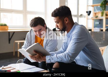 Junger glücklicher Mann mit Down-Syndrom und sein Tutor, der in der Schule drinnen studiert. Stockfoto