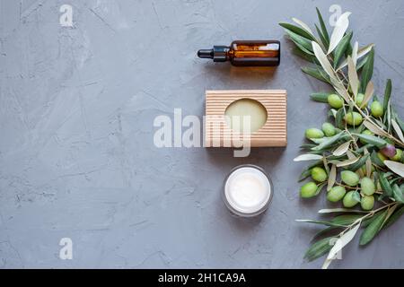 Konzept von natürlichen hausgemachten Zutaten in der Kosmetik für die Hautpflege. Bio-Olivenöl in Glasflasche, handgefertigtes Seifenstück und Creme mit Olivenzweigen. Ruhe und Entspannung. Grauer Hintergrund. Stockfoto