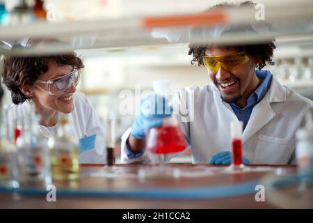 Junge Kollegen in Schutzkleidung sind gut gelaunt, während sie im Universitätslabor in entspannter Atmosphäre mit bunten Chemikalien arbeiten. Scie Stockfoto