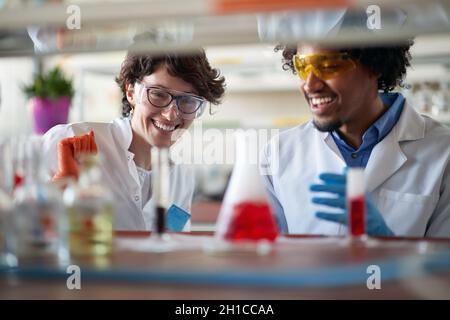 Junge Kollegen in Schutzkleidung analysieren farbenfrohe Chemikalien in entspannter Atmosphäre im Universitätslabor. Wissenschaft, Chemie, Labor, Menschen Stockfoto