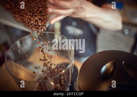 Nahaufnahme der Hände, die einen Behälter halten und duftende und aromatische Kaffeebohnen in einen Mahlwerk-Apparat gießen. Kaffee, Getränke, Produktion Stockfoto