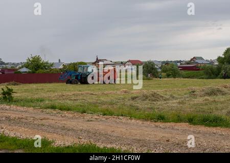 Nischni Nowgorod, Russland - 1. Juli 2020: Blauer Traktor sammelt Heu in Ballen, Heuernte am Ende des Sommers. Dorfbewohner treffen sich, während sie Heu sammeln Stockfoto