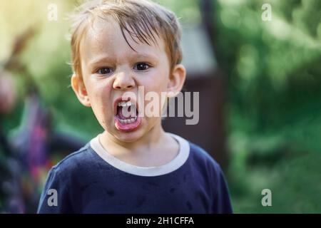 Wütender kleiner Junge mit nassem Haar in blauem T-Shirt schreit auf verschwommenem Hintergrund im grünen Stadtpark aus der Nähe Stockfoto