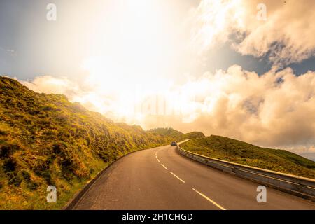 Auto auf einer kurvenverlaufenen Straße mit Sonnenuntergang über der Straße in der Nähe von Lagoa do Fogo mit einer wunderschönen Naturlandschaft. Insel São Miguel auf den Azoren, Portugal. Stockfoto