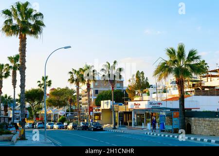 Paphos, ZYPERN - 13. FEBRUAR 2019: Stadtbild der Paphos Innenstadtstraße mit Palmen, Geschäften und geparkten Autos in Abendlicht Stockfoto